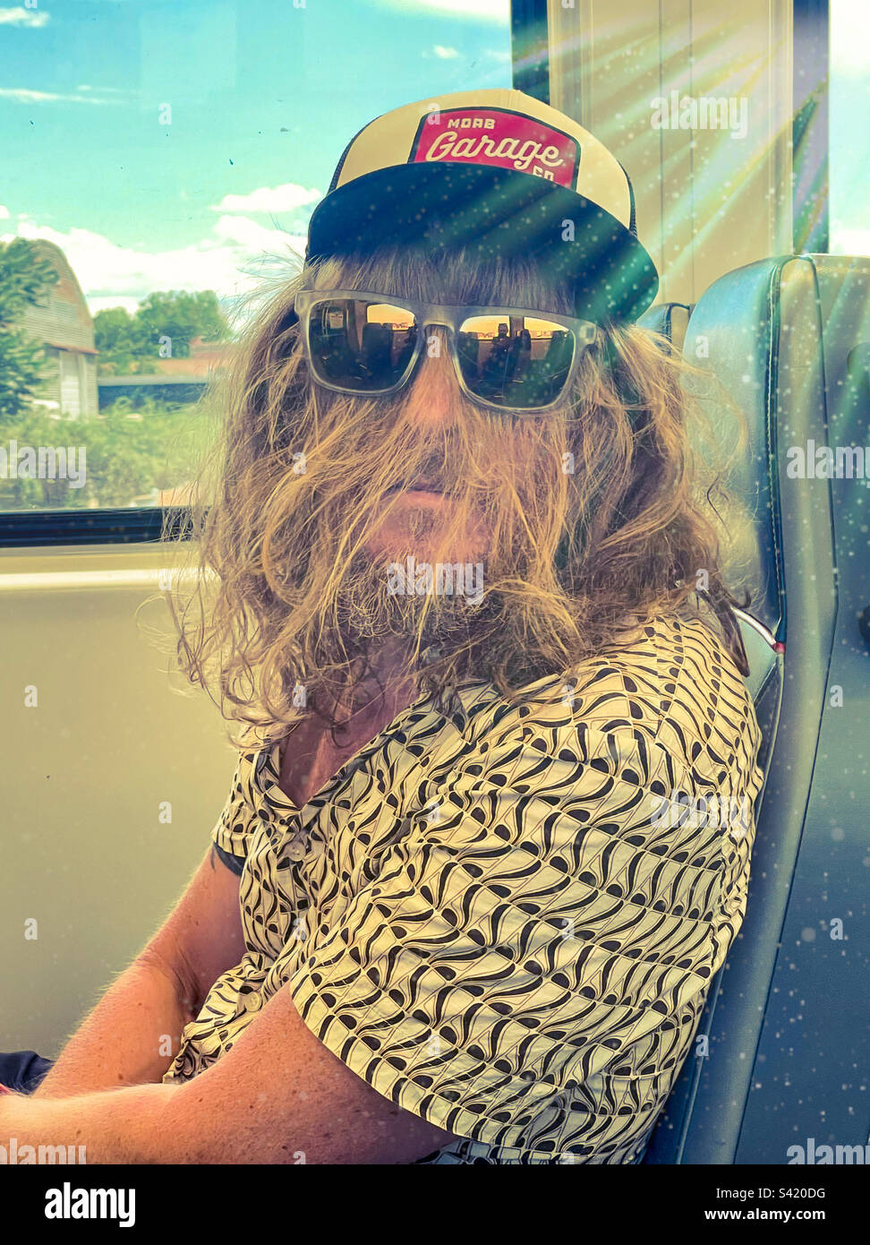 Ein Mann mit langen Haaren und einer Sonnenbrille, der im Zug rummacht - Smartphone-aufgenommenes Stockfoto