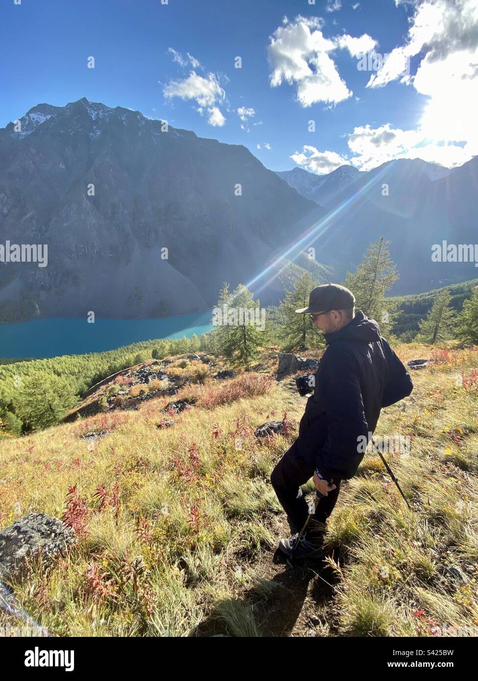Ein männlicher Touristenfotograf geht den Berg hinunter zu den Bäumen in der Nähe eines türkisfarbenen Sees unter einem Felsen mit Sonnenlicht. - Smartphone-aufgenommenes Stockfoto