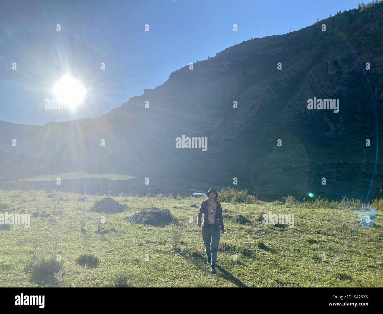 Ein Mädchen geht durch ein helles Feld in den Sonnenstrahlen hinter einem Berg im Schatten des Altai in Sibirien. - Smartphone-aufgenommenes Stockfoto