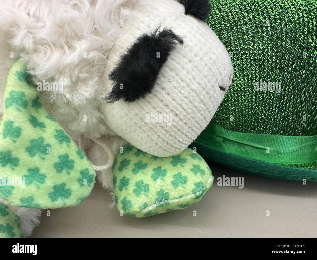 Lamm mit St. Patrick's Day Flair. Plüschtier mit Shamrocks an den Ohren. Und der schimmernde, grüne Hut daneben. - Smartphone-aufgenommenes Stockfoto