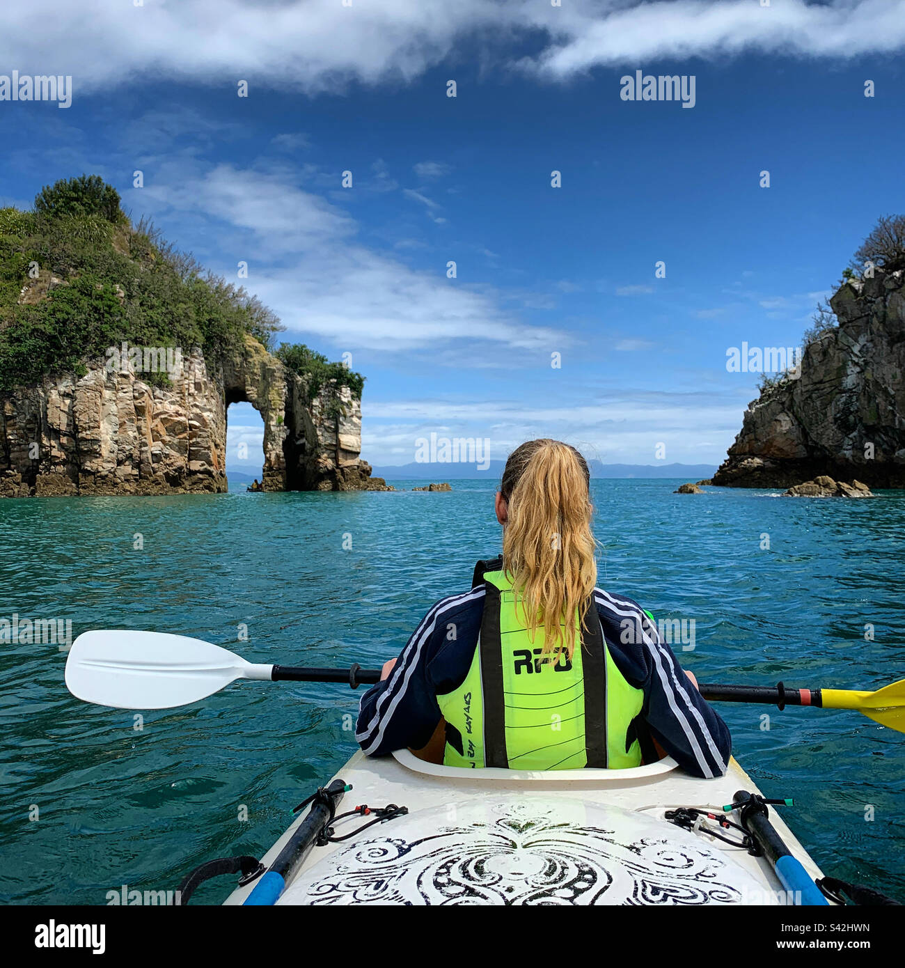 Ein junges Mädchen im Teenageralter in einem Kajak, das auf einen Bogentunnel in einer Klippe in der Golden Bay Gegend von South Island Neuseeland schaut Stockfoto