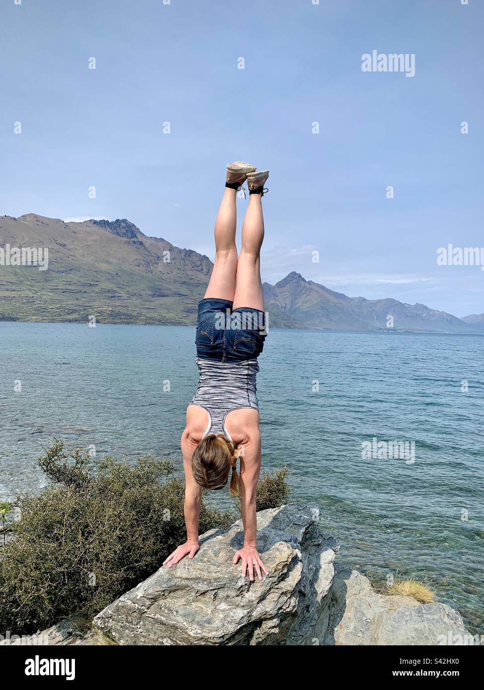 Eine sportliche Frau, die einen Handstand am Lake Wakatipu auf der Halbinsel Kelvin Heights bei Queenstown auf der Südinsel Neuseeland macht - Smartphone-aufgenommenes Stockfoto