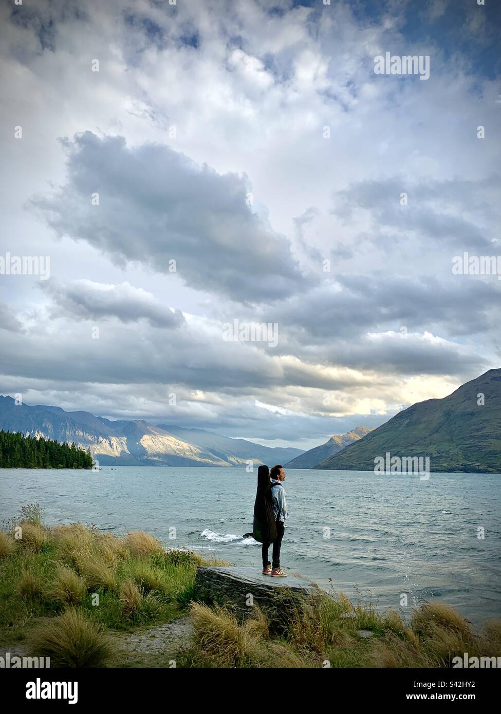 Ein Musiker mit einer Gitarre auf dem Rücken, der über den Lake Wakatipu in der Nähe von Queenstown auf der Südinsel Neuseeland blickt - Smartphone-aufgenommenes Stockfoto