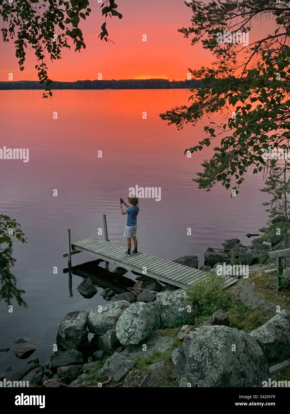 Ein Teenager macht mit seinem Handy ein Foto von Mitternachtssonne und einem blutroten Himmel über einem Seepier in einem Sommerhaus in der finnischen Kajaani-Region Stockfoto