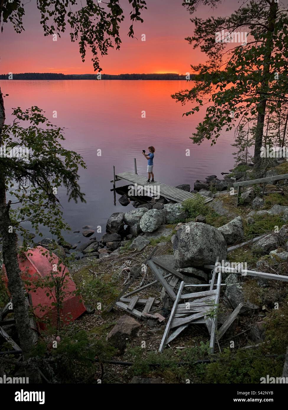 Ein Teenager macht mit seinem Handy ein Foto von Mitternachtssonne und einem blutroten Himmel über einem Seepier in einem Sommerhaus in der finnischen Kajaani-Region Stockfoto