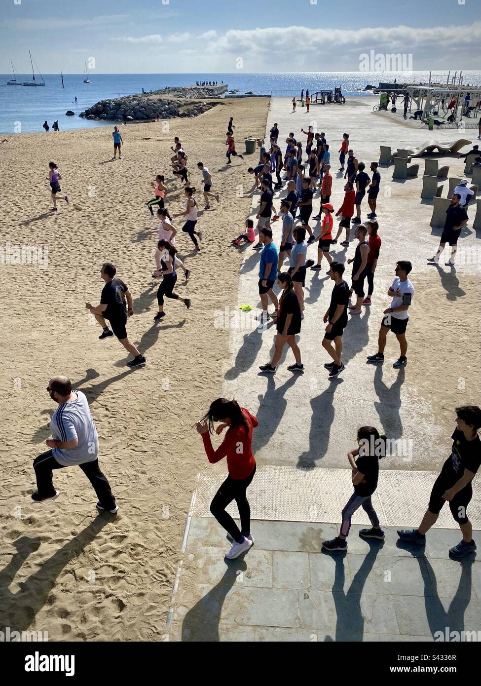 Eine Gruppe von Menschen jeden Alters, die morgens Intervalle und Sprints am geschäftigen Stadtstrand von Barceloneta in Barcelona, Spanien, machen - Smartphone-aufgenommenes Stockfoto