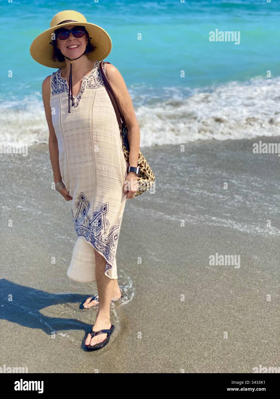 Eine Frau, die am Strand spaziert, einen Strohhut mit breiter Krempe und eine Sonnenbrille trägt. Stockfoto