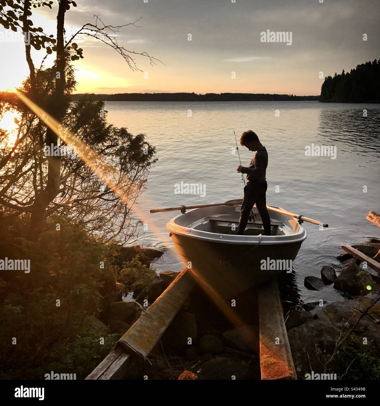 Ein kleiner Junge steht in einem Boot mit einer Angelrute bei Sonnenuntergang und bereitet sich darauf vor, in der Mitternachtssonne an einem Sommersee in der finnischen Kajaani-Region angeln zu gehen Stockfoto