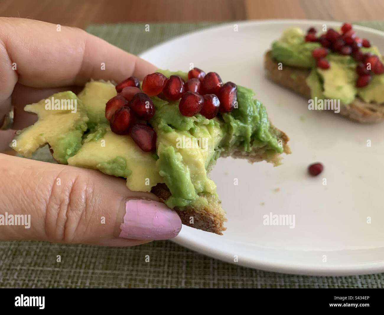 Nahaufnahme einer Frau, die ein halb gefressenes Stück Sauerteig-Avocado-Toast mit Granatäpfeln in der Hand hält - Smartphone-aufgenommenes Stockfoto