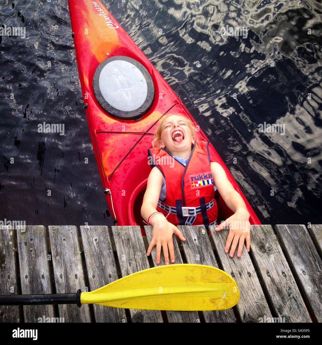 Ein junges Mädchen in einer Schwimmweste, das in einem Kajak lacht, während es während eines arktischen Sommers unter einem Pier an einem See in der abgelegenen Kajaani-Region Finnlands untergeht Stockfoto