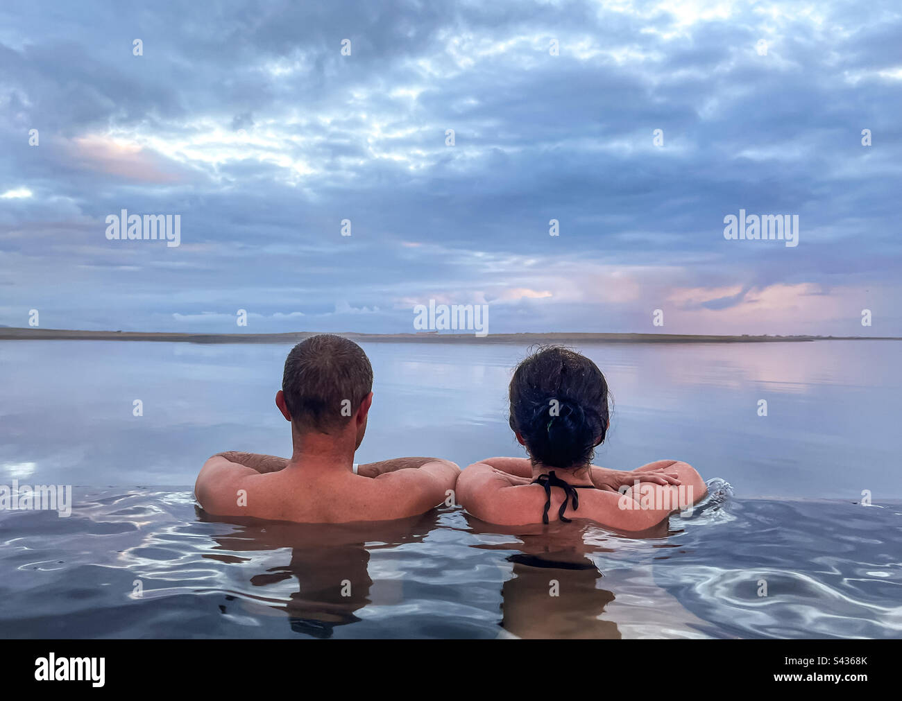 Ein Paar von hinten in einem Infinity Hot Pool mit Blick auf den Ozean bei Sonnenuntergang, geothermischer Pool in Island, romantische Szene Stockfoto