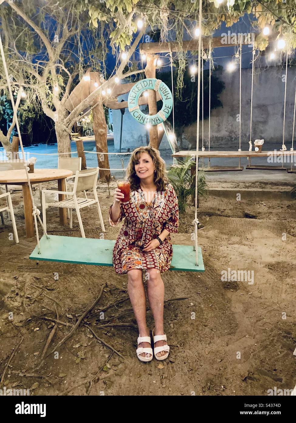 Eine Frau mit einem Drink in der Hand sitzt auf einer Schaukel im Nereida Cantina in Holbox. - Smartphone-aufgenommenes Stockfoto