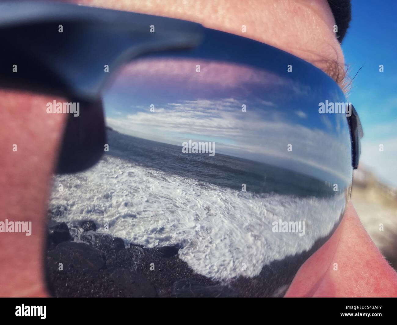 Das Meer spiegelt sich in der Sonnenbrille eines Mannes, der am Strand steht - Smartphone-aufgenommenes Stockfoto