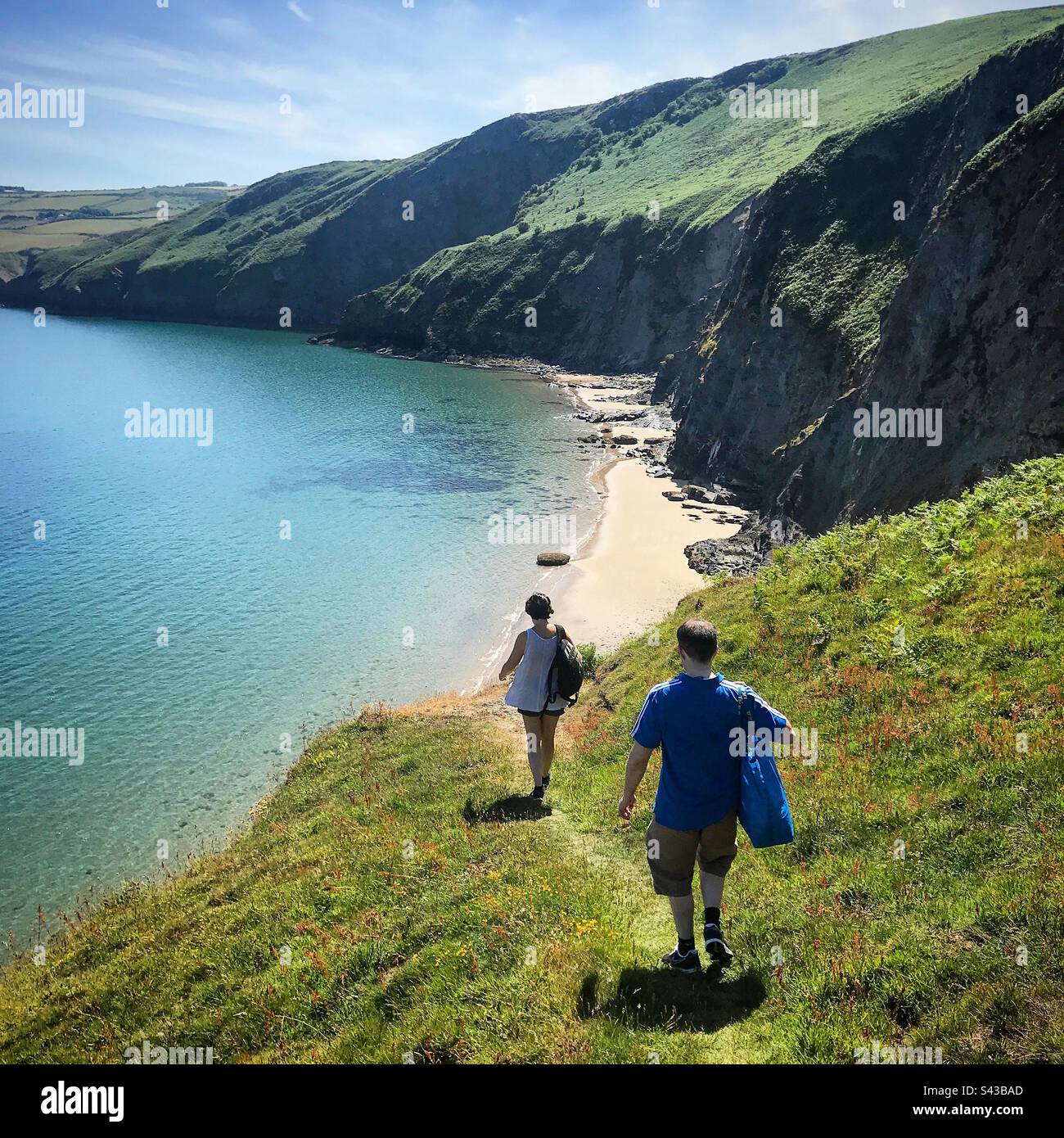 Zwei Touristen steigen hinab zu einem romantischen, geheimen, versteckten Strand unter einer Klippe auf einem dramatischen Abschnitt des beliebten Ceredigion Coast Path in der Nähe von Llangrannog in West Wales Stockfoto