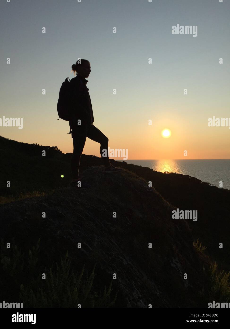 Eine Frau in Silhouette bei Sonnenuntergang, die über der Irischen See auf einem dramatischen Abschnitt des beliebten Ceredigion Coast Path in der Nähe von Llangrannog in Westwales spaziert Stockfoto