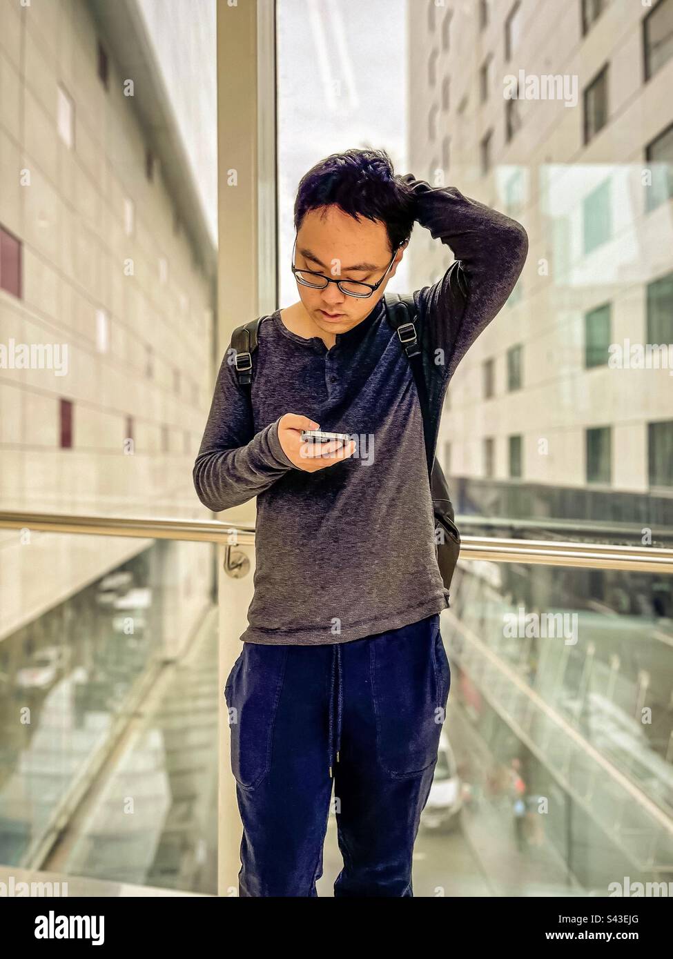 Junger Asiate in Brille mit Handy gegen Fensterblick auf Straße und Gebäude in der Stadt. - Smartphone-aufgenommenes Stockfoto