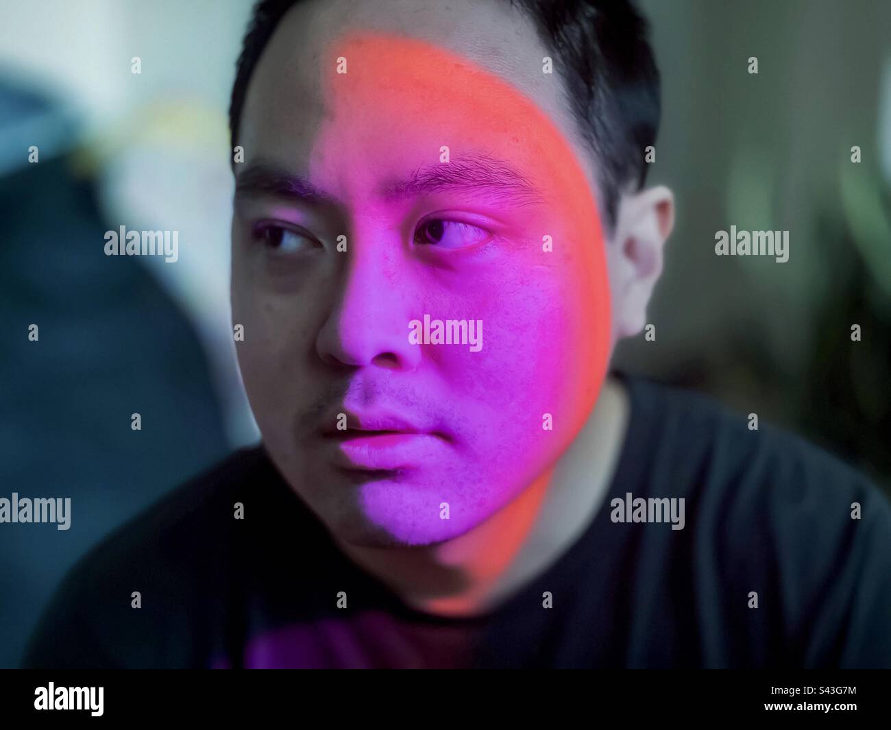Nahaufnahme eines jungen Asiaten mit farbigem LED-Licht, das die Hälfte seines Gesichts vor unscharfem Hintergrund beleuchtet. - Smartphone-aufgenommenes Stockfoto