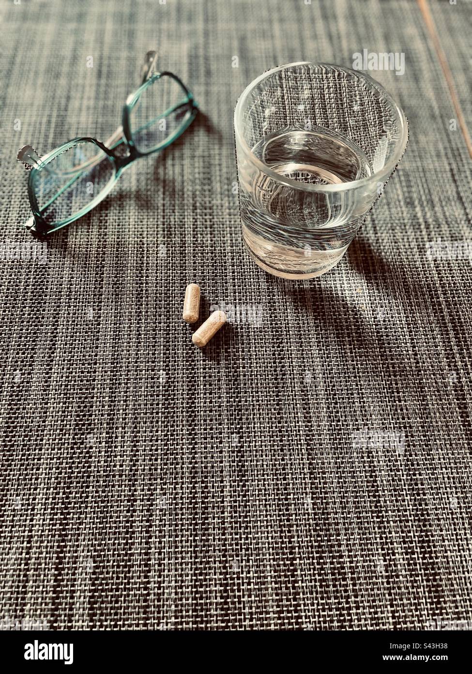 Morgendosis, zwei Kapseln auf einem strukturierten Hintergrund mit einem Glas Wasser und einem Paar Gläser auf der Seite Stockfoto