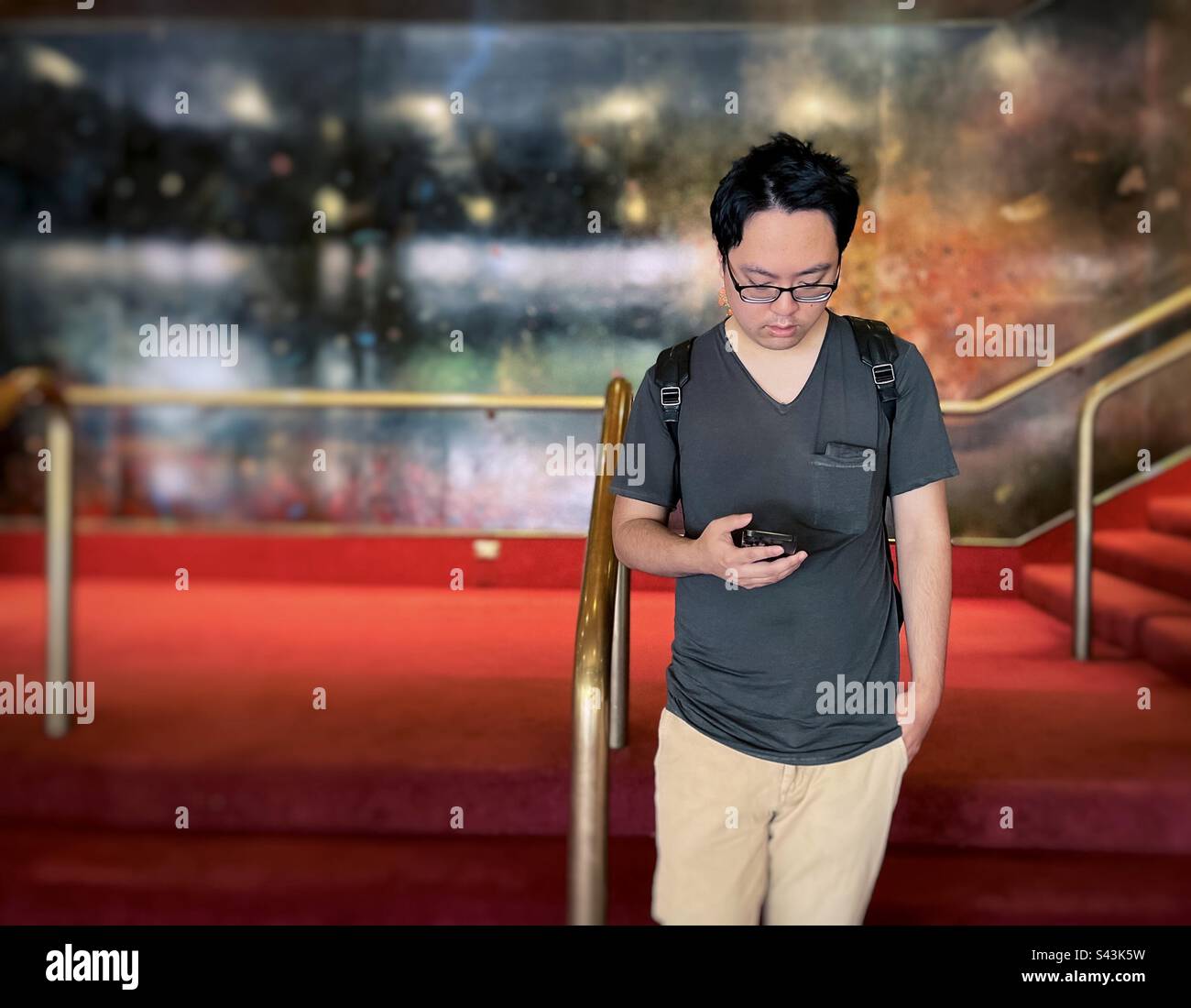 Ein junger asiatischer Mann, der sein Smartphone auf der Treppe gegen Geländer und Wand benutzt. - Smartphone-aufgenommenes Stockfoto