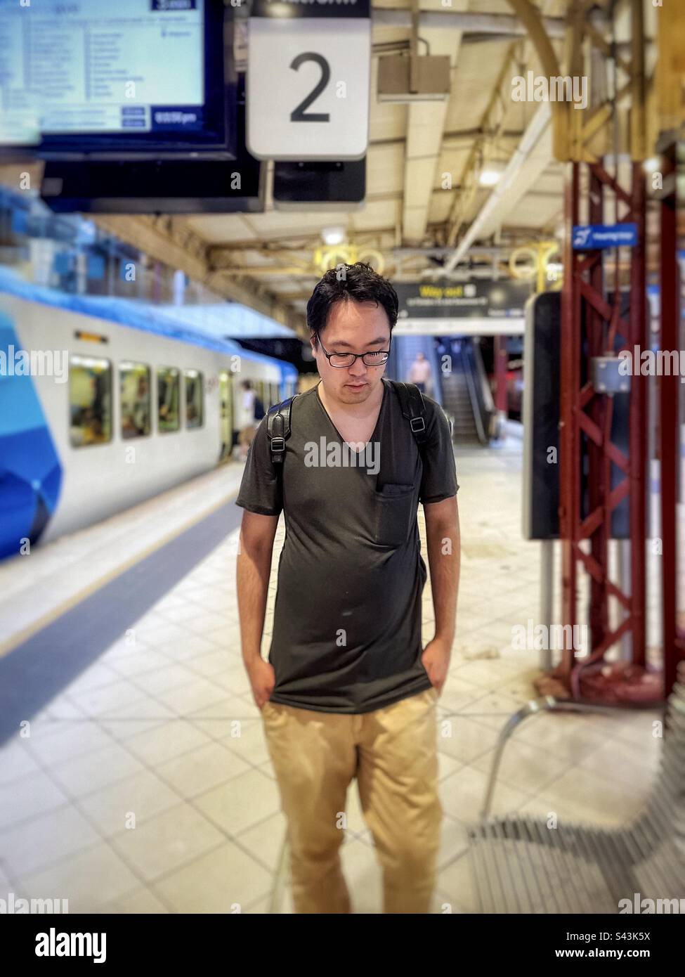 Ein junger asiatischer Mann mit Brille steht am Bahnhofsplatz in Melbourne, Victoria, Australien, selektiver Fokus. - Smartphone-aufgenommenes Stockfoto