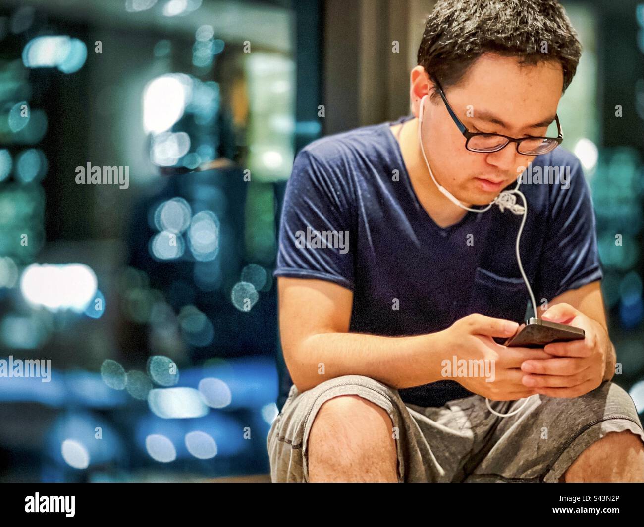 Junger asiatischer Mann mit Kopfhörern und Brillen, der Handy benutzt, gegen Unschärfe-Reflexionen im Fenster in der Nacht. - Smartphone-aufgenommenes Stockfoto