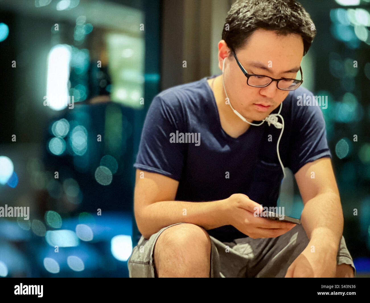 Junger asiatischer Mann mit Kopfhörern und Brillen, der sein Smartphone gegen Unschärfe-Reflexionen in Fenstern in der Nacht benutzt. - Smartphone-aufgenommenes Stockfoto