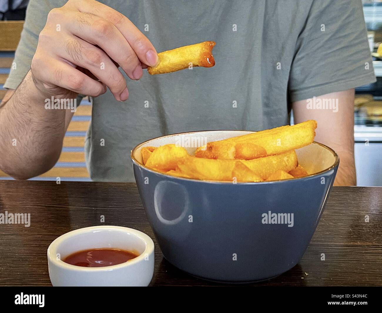 In der Mitte der Hand eines Mannes, der halb gegrillte pommes frites in Tomatensoße über einer Schüssel mit Pommes frites und Tomatensoße in einer Schüssel auf dem Tisch hält. Tröstungsessen. Sehnsucht. - Smartphone-aufgenommenes Stockfoto