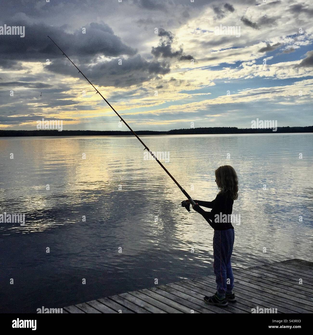 Ein junges 8-jähriges Mädchen steht auf einem Pier mit einer Angelrute bei Sonnenuntergang in der Mitternachtssonne in einem Sommerhaus in der finnischen Kajaani-Region Stockfoto