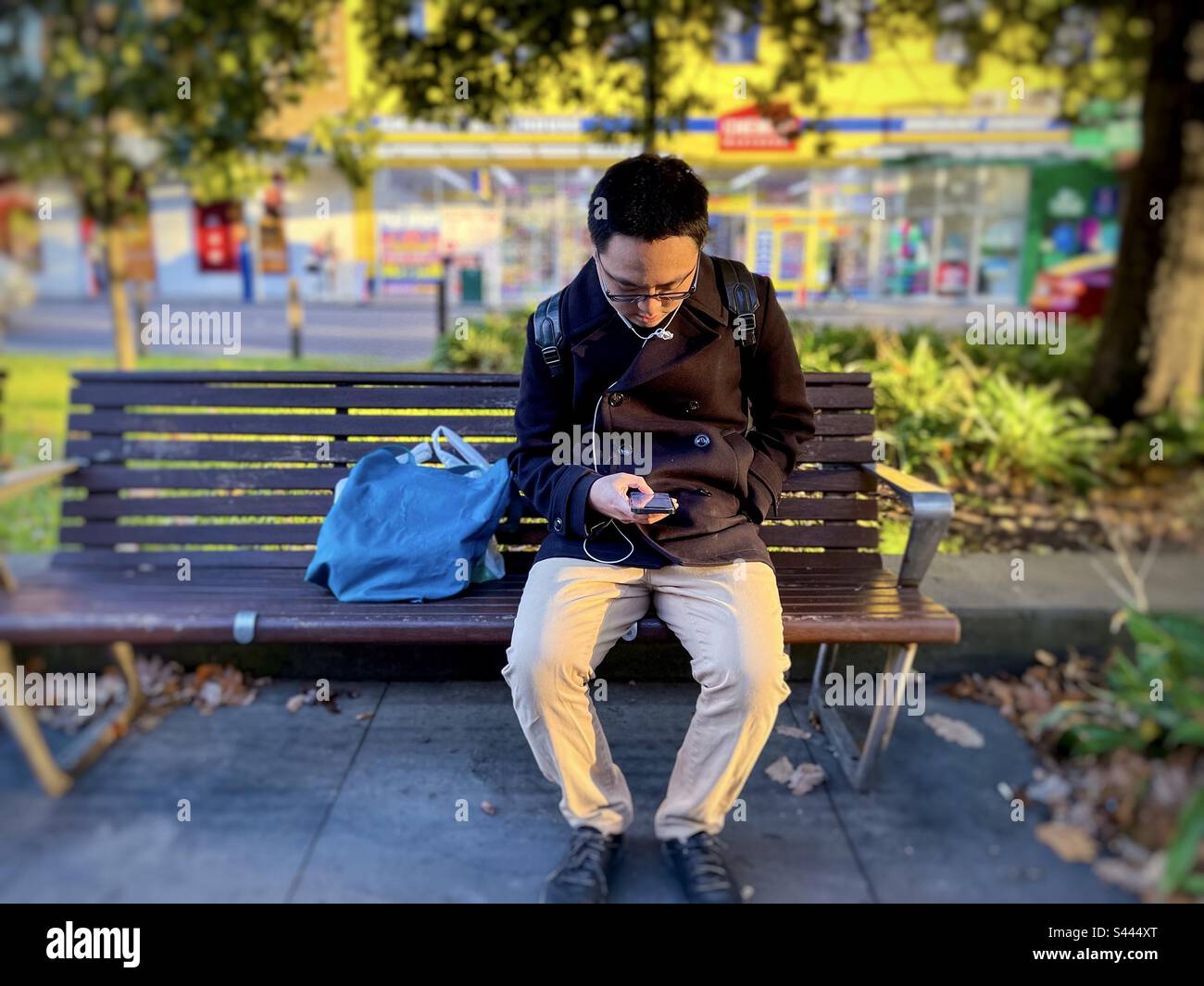 Ein junger asiatischer Mann, der mit dem Handy telefoniert, während er auf einer Parkbank an Bäumen, Gras, Straßen und Geschäften sitzt. Konzentrieren Sie sich auf den Vordergrund. Sonnenuntergang. - Smartphone-aufgenommenes Stockfoto