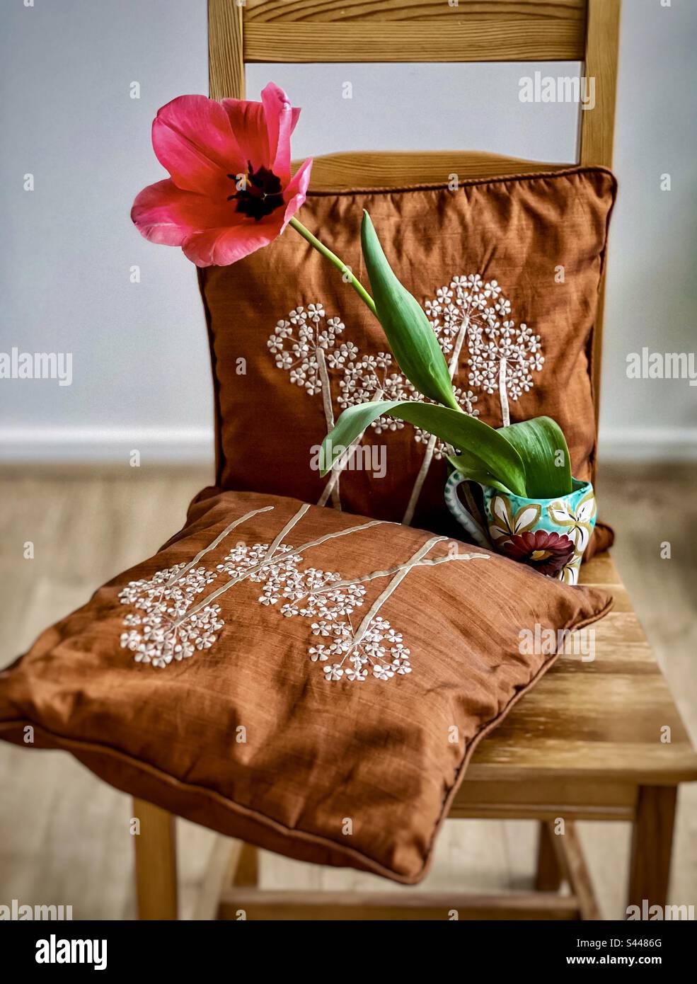 Eine pinkfarbene Tulpenblume in Vase mit gestickten Kissen auf einem Holzstuhl im Wohnzimmer. Böhmischer rustikaler Stil. Frühjahrsthema. - Smartphone-aufgenommenes Stockfoto