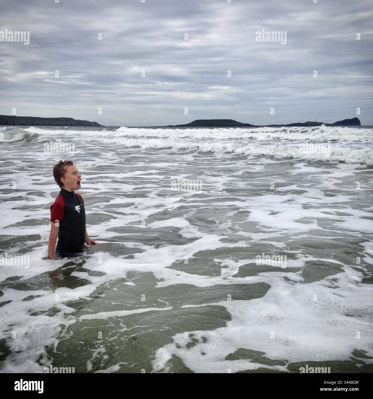 Ein neunjähriger Junge springt in das kalte Wasser und trotzt den großen Wellen in einem Neoprenanzug in der Irischen See am Rhossili Llangennith Beach in West Wales Stockfoto