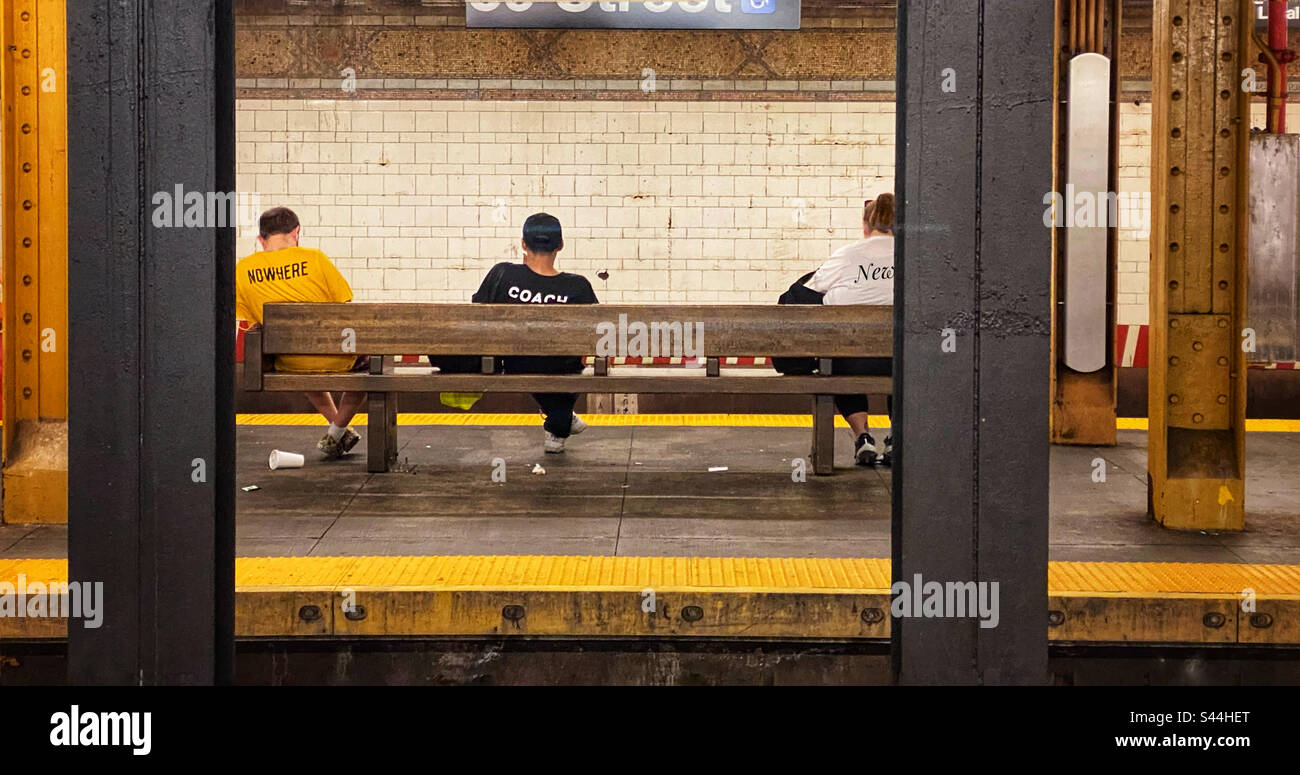 Drei Leute mit Wörtern auf ihren T-Shirts, die auf einer Bank sitzen und in einer U-Bahn-Station auf einen Zug warten Stockfoto