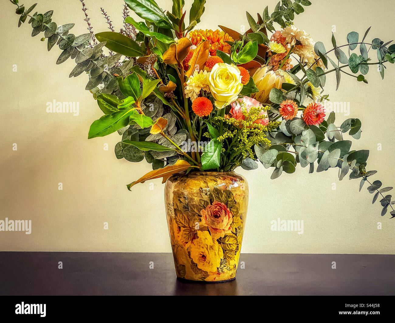 Blumenstrauß aus frischen Blumen und Blättern in einer Vase mit Blumenmuster und Dekoupation im Vintage-Stil auf einem schwarzen Tisch an der weißen Wand. Art. Vintage-Style. Blumendesign. Stockfoto