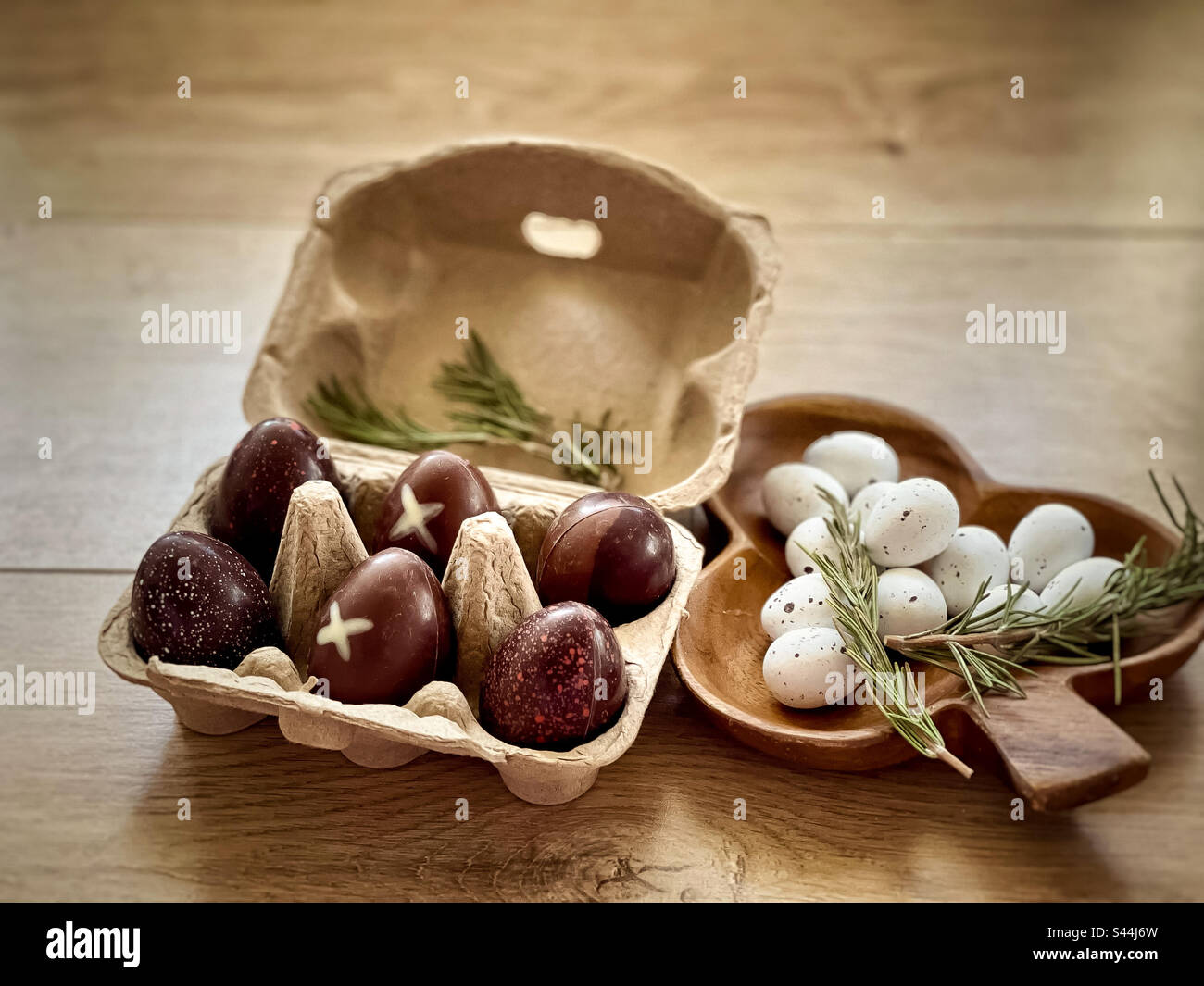 Blick aus einem hohen Winkel auf Schokoladeneier in Eierkarton aus Pappe und praline Ostereier in dekorativen Holzbehältern mit Zweigen Rosmarin auf Holztisch. Rustikaler Stil. Ostern. Feier. - Smartphone-aufgenommenes Stockfoto