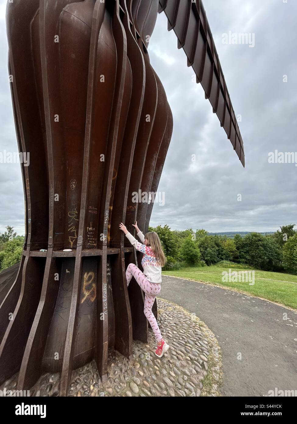 Ein 8-jähriges Mädchen klettert den Fuß der Engel-des-Nordens-Statue in Newcastle hoch - Smartphone-aufgenommenes Stockfoto
