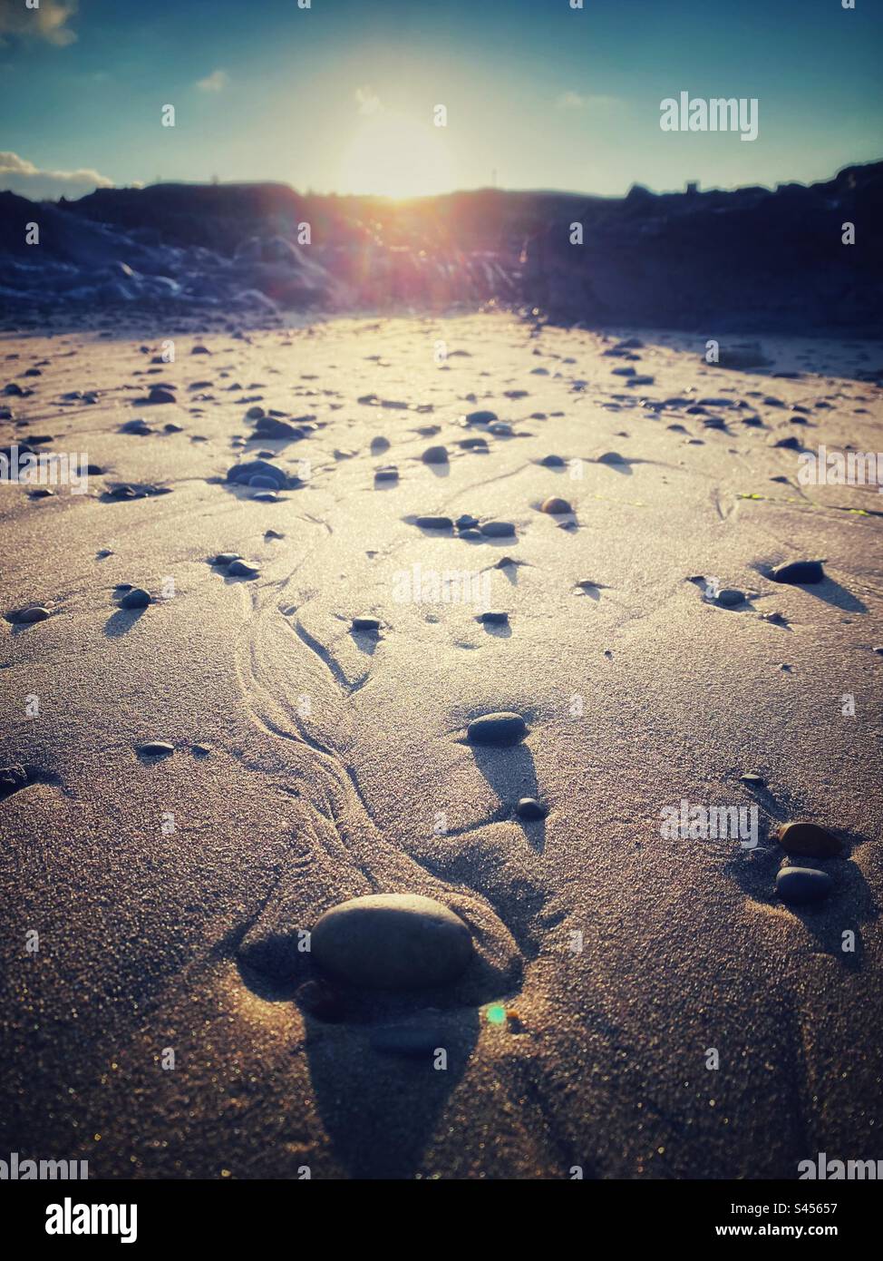 Ein gefiltertes Foto mit geringer Tiefenschärfe von Kieselsteinen an einem Strand bei Sonnenaufgang, mit Rillen auf dem Sand, die vom Meer verursacht werden. Wunderschöne Strandszene an der Küste - Smartphone-aufgenommenes Stockfoto