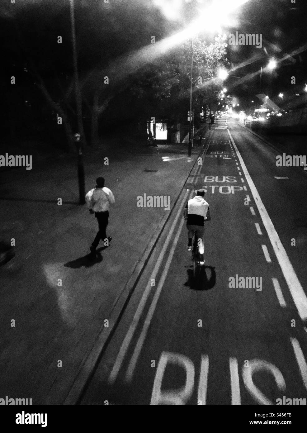 Ein Radfahrer fährt mit seinem Fahrrad auf der Busspur, während ein Mann nachts in London in Richtung einer Bushaltestelle auf dem Bürgersteig rennt - Smartphone-aufgenommenes Stockfoto