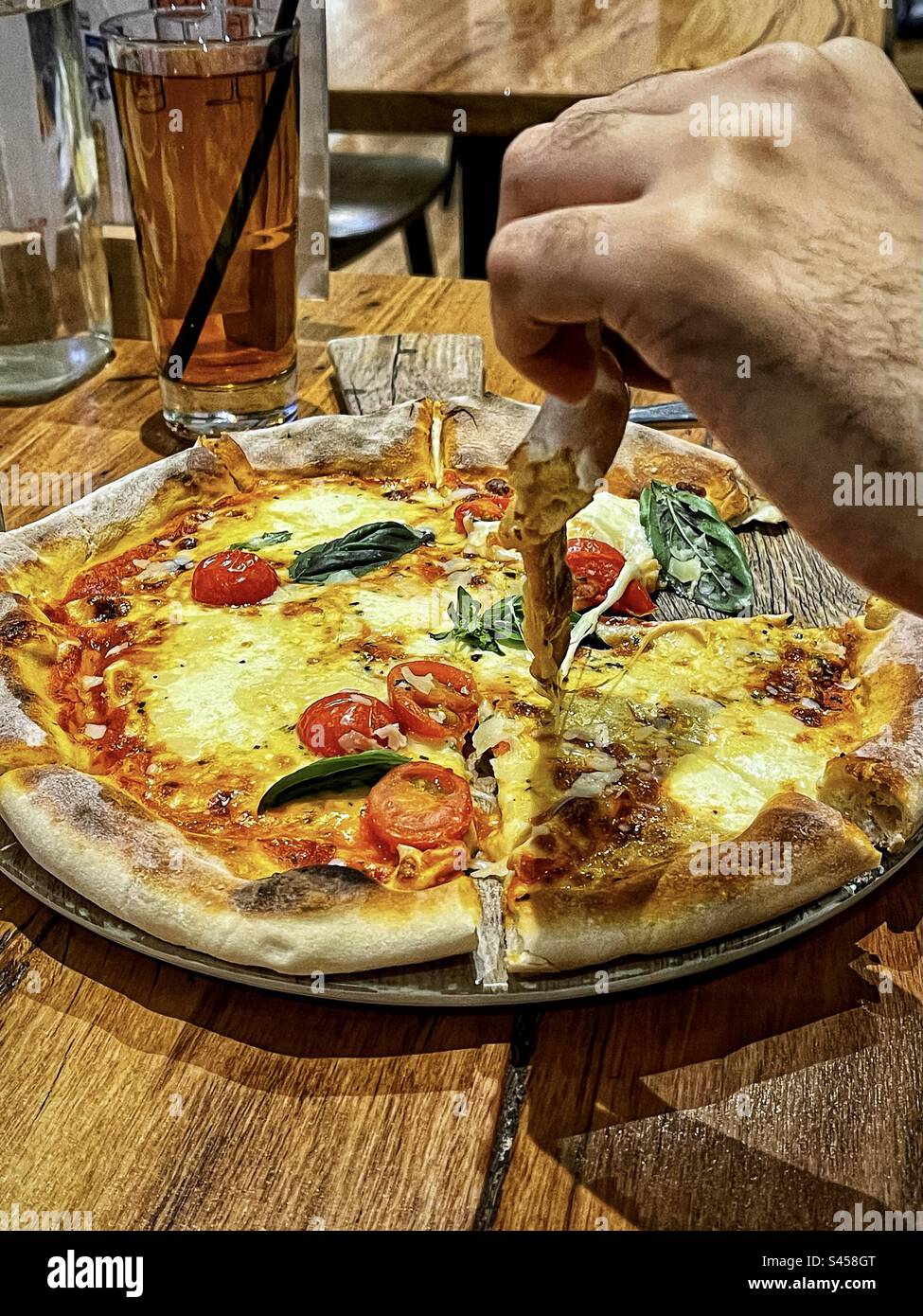 Nahaufnahme einer Hand, die ein Stück Pizza von Pizza Magherita auf dem Servierbrett auf dem Tisch abholt. - Smartphone-aufgenommenes Stockfoto
