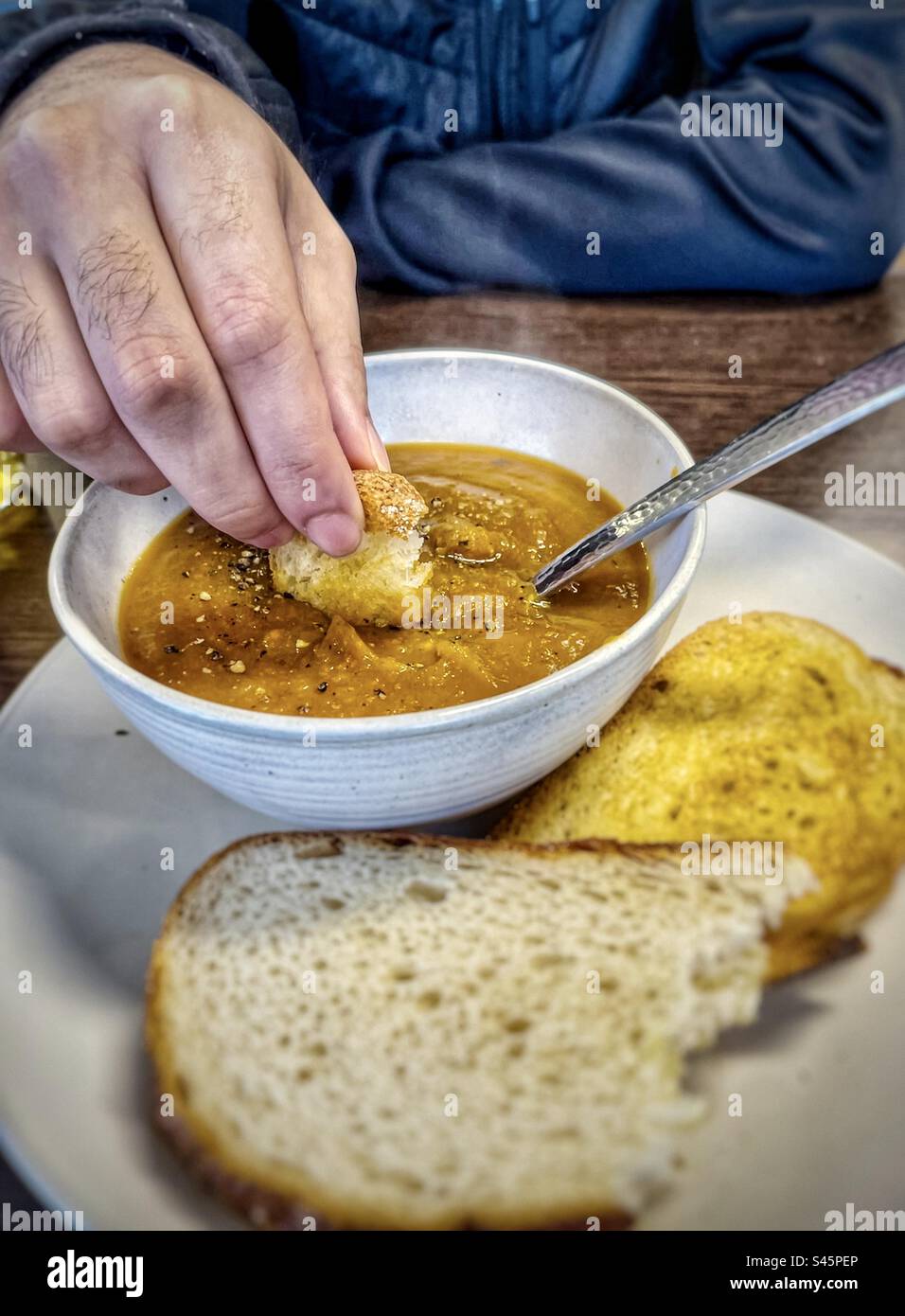 In der Mitte des Mannes, der Brot in eine Schüssel dampfender, heißer Kürbissuppe mit Buttertoast auf dem Teller eintaucht. - Smartphone-aufgenommenes Stockfoto