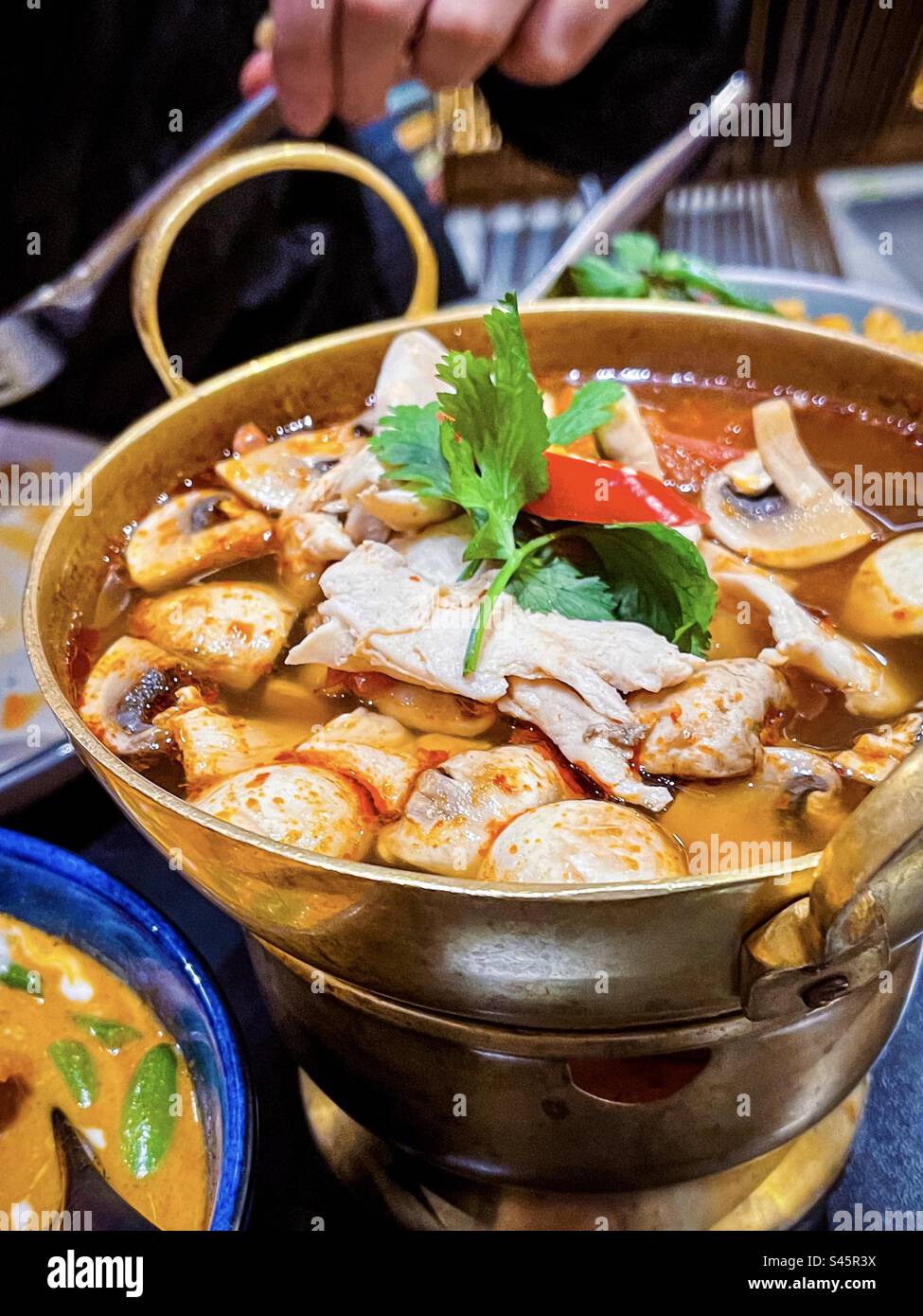 Blick aus einem hohen Winkel auf die scharfe thailändische Tom Yum Suppe mit Pilzen, Hühnchen und Kräutern im Hot Topf, mit Hand, die im Hintergrund einen Löffel hält. - Smartphone-aufgenommenes Stockfoto