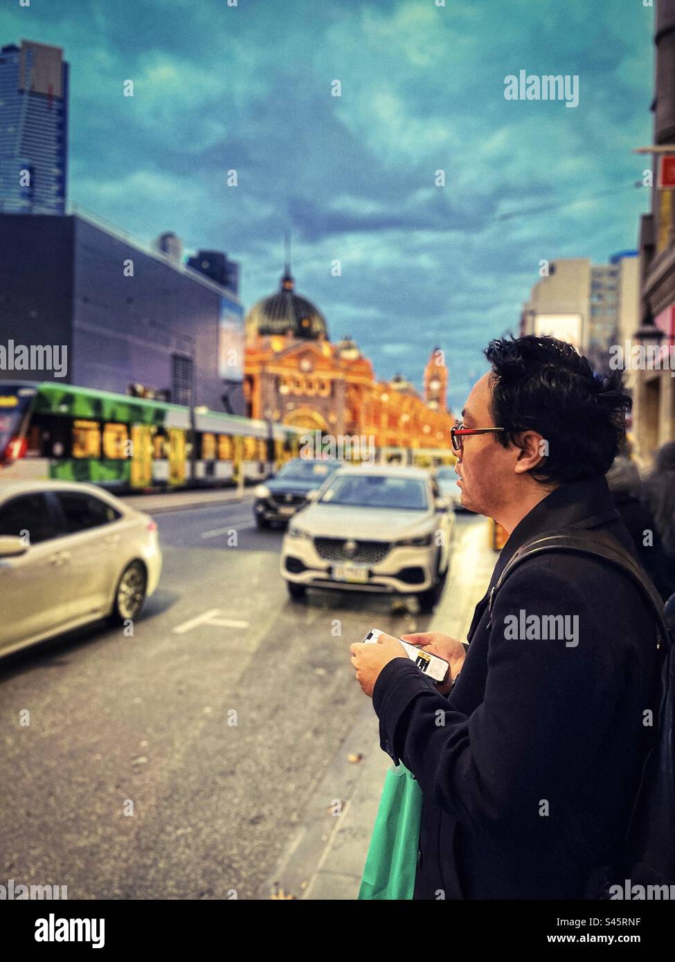 Junger Mann, der ein Mobiltelefon benutzt, während er auf einem Bürgersteig neben einer geschäftigen Straße in Melbourne CBD, Victoria, Australien steht. Guten Abend. Konzentrieren Sie sich auf den Vordergrund. - Smartphone-aufgenommenes Stockfoto