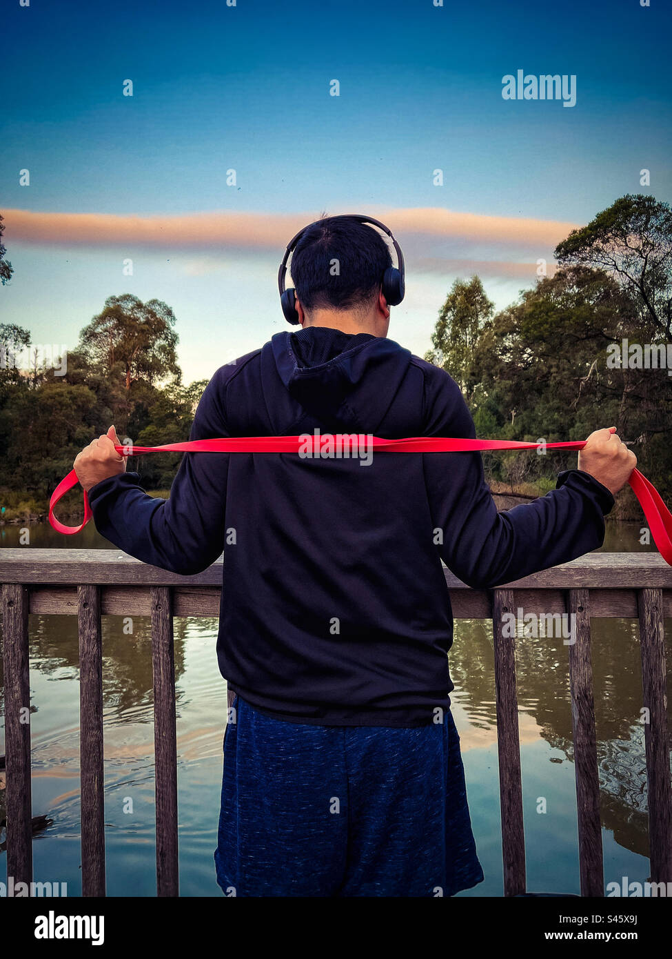 Rückansicht eines jungen Mannes mit Kopfhörern, der mit einem Widerstandsband im Park trainiert, gegen die rosa Wolke am Himmel, an Bäumen und am See bei Sonnenuntergang. Outdoor-Training. Ruhige Szene. - Smartphone-aufgenommenes Stockfoto