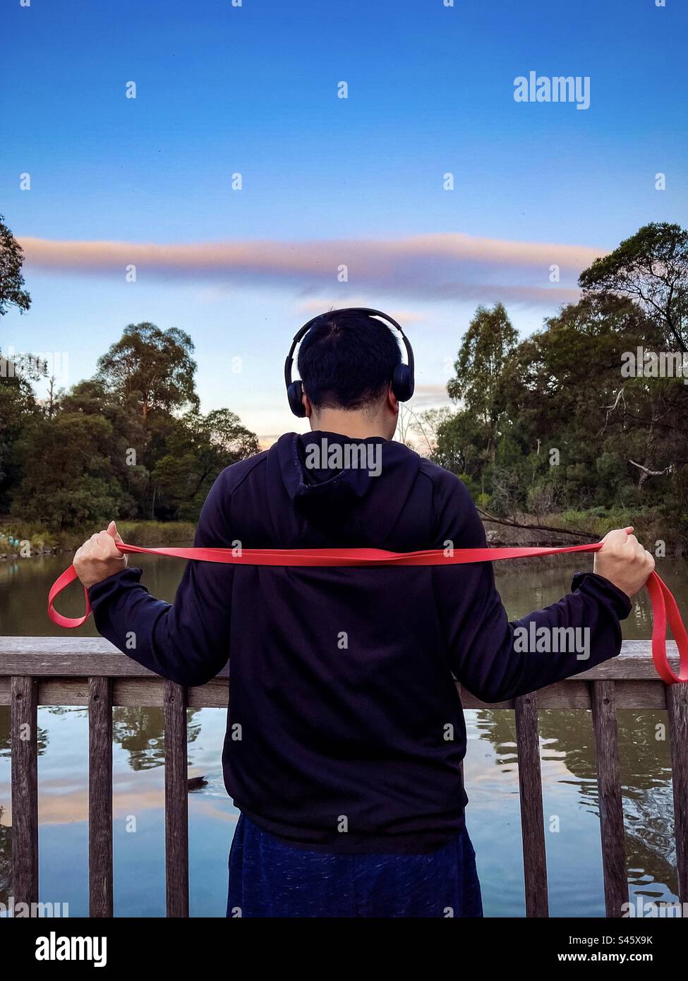 Rückansicht eines jungen Mannes mit Kopfhörern, der mit einem Widerstandsband im Park vor rosa Wolken am Himmel, Bäumen und See bei Sonnenuntergang trainiert. Outdoor-Training. Ruhige Szene. - Smartphone-aufgenommenes Stockfoto