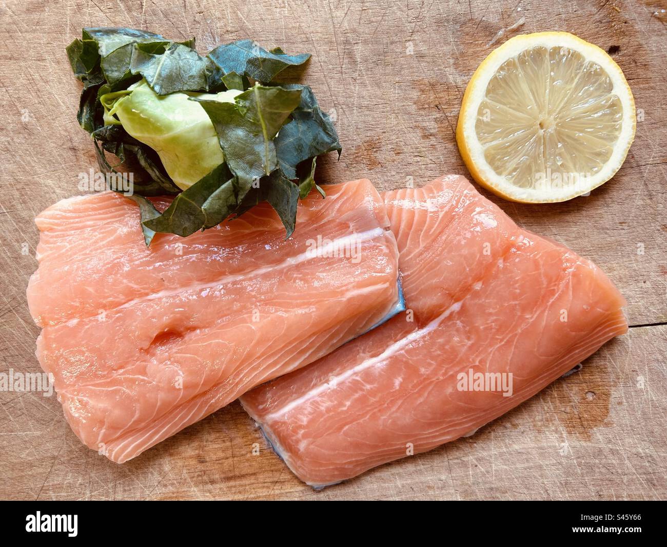 Frische, rohe, nicht gekochte, nahrhafte Lachsschwänze, Zitronenscheiben und gehackter grüner Kohl und ein Küchenmesser mit Holzgriff auf einem Schneidebrett. Fertig zum Kochen. - Smartphone-aufgenommenes Stockfoto