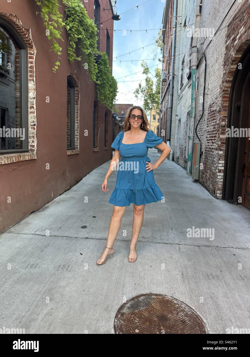 Eine Frau, die eine Sonnenbrille und ein blaues Kleid trägt und in einer Gasse in der Innenstadt posiert. Stockfoto