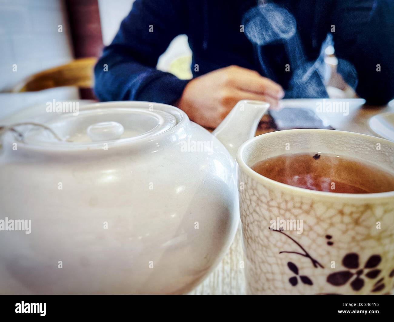 Nahaufnahme von Dampf, der sich von einer Tasse heißen chinesischen Tee neben einer weißen Teekanne gegen die Mitte eines Mannes dreht, der ein Mobiltelefon am Tisch im Café benutzt. - Smartphone-aufgenommenes Stockfoto