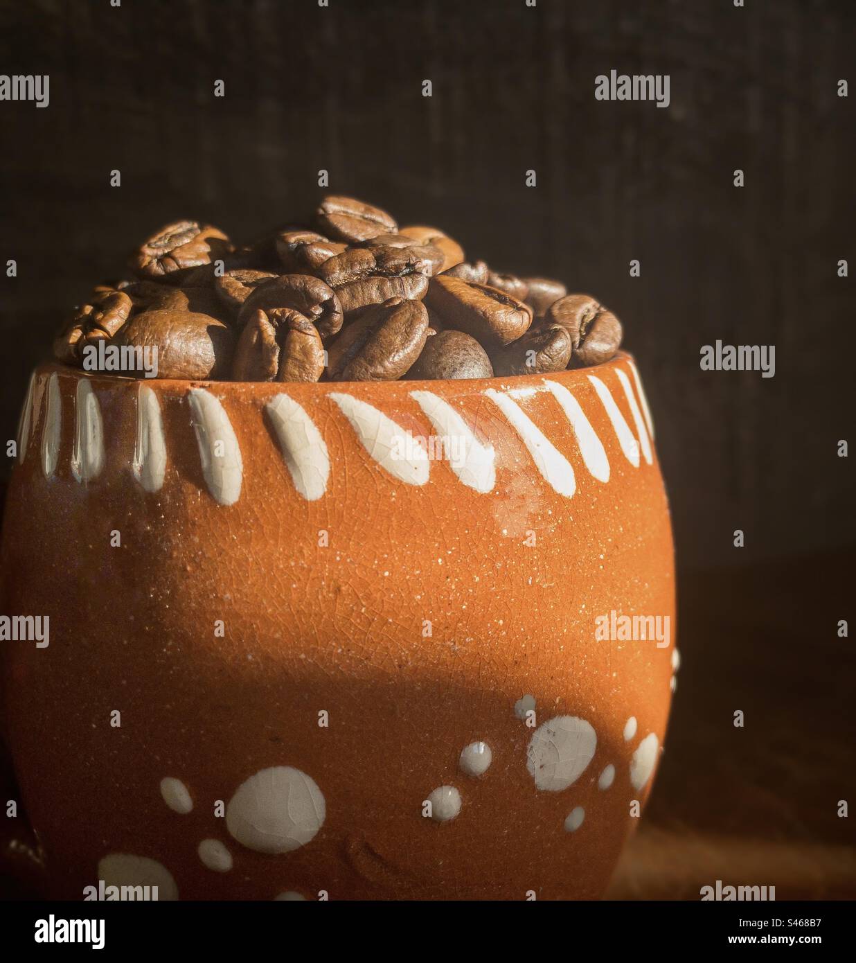 Eine Terrakotta-Tasse gefüllt mit Kaffeebohnen, die sich in einem Schacht von Sonnenlicht verfangen hat - Smartphone-aufgenommenes Stockfoto