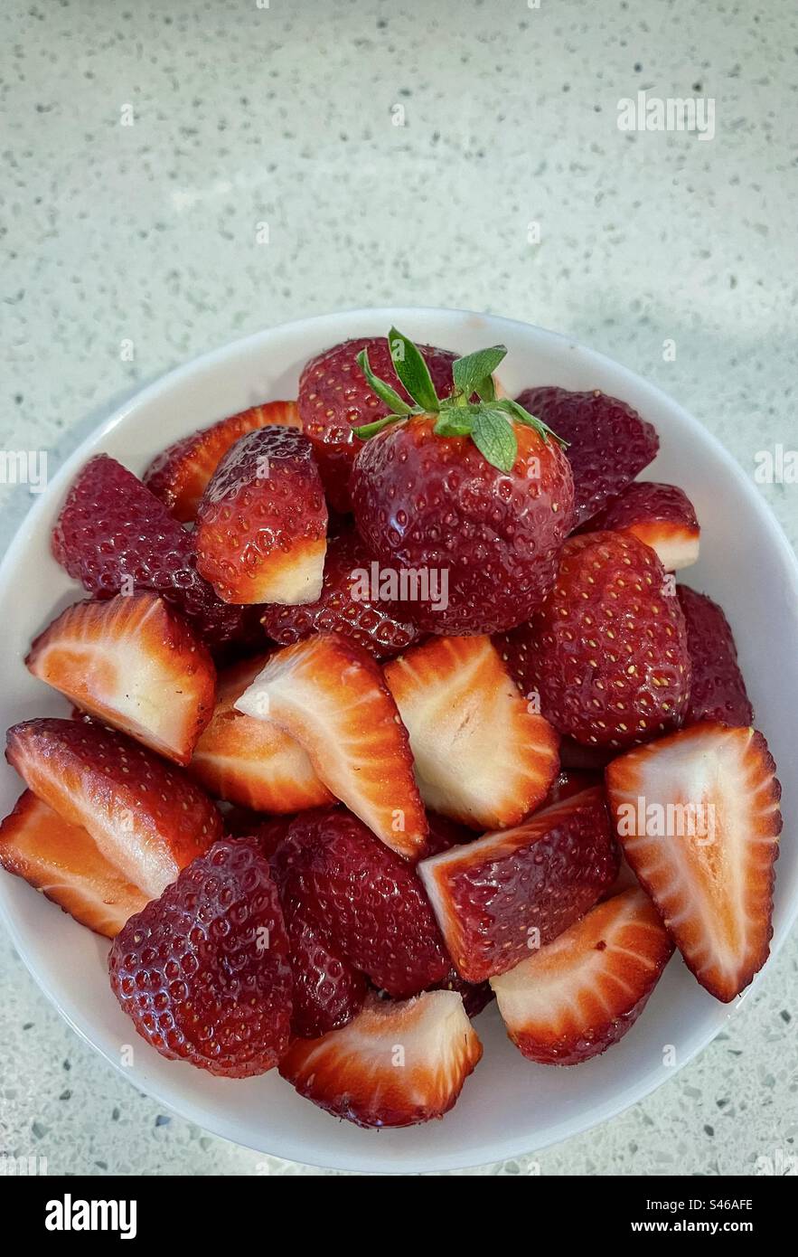 Blick aus einem hohen Winkel auf eine weiße Schüssel mit geschnittenen, organischen, frischen Erdbeeren mit einer einzigen ganzen Erdbeere oben auf einer weißen Arbeitsfläche. - Smartphone-aufgenommenes Stockfoto