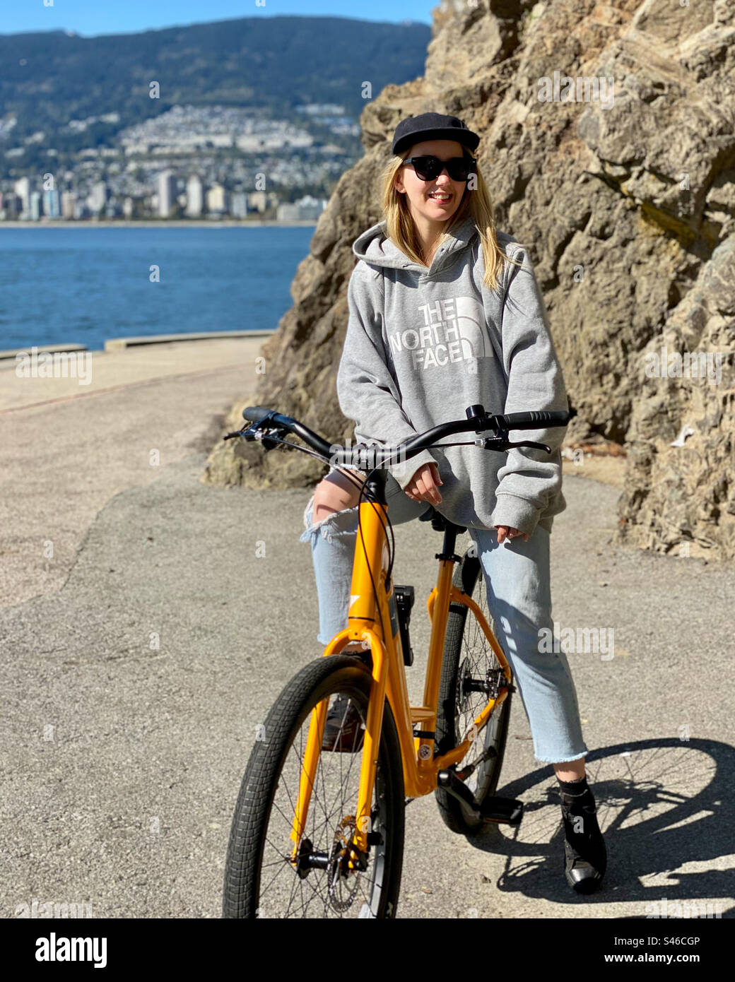 Junge blonde Hipster-Frau sitzt auf einem Fahrrad auf einem Strandweg, während sie nach rechts von der Kamera schaut. - Smartphone-aufgenommenes Stockfoto