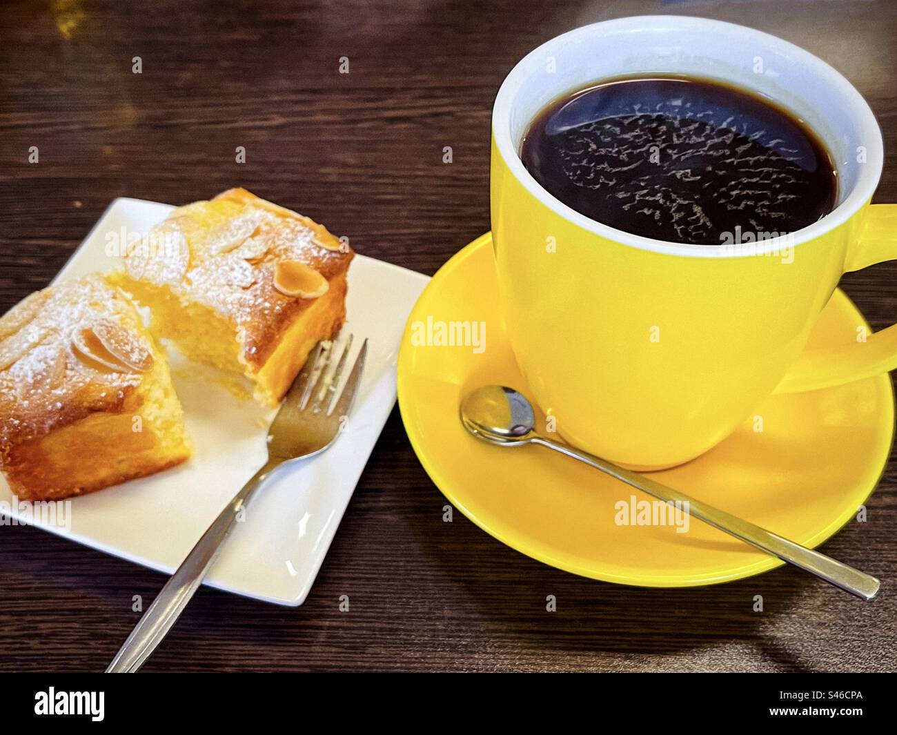 Kaffeepause. Ein langes Schwarz in einer gelben Tasse und Untertasse mit einer Scheibe glutenfreiem Orangenkuchen auf einem Teller auf einem Holztisch. Stockfoto